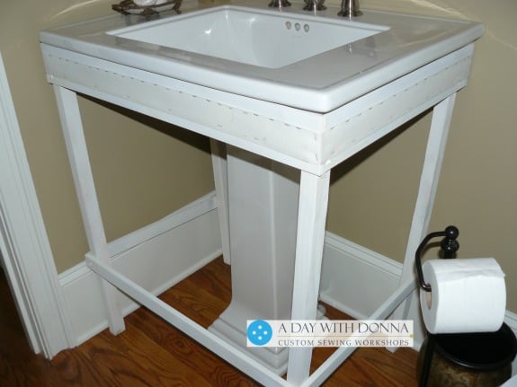 Make a frame from lumber to fit under the lip of the sink.   Attach Velcro hook along the sides and front of the frame.  This will hold your skirt to the frame.